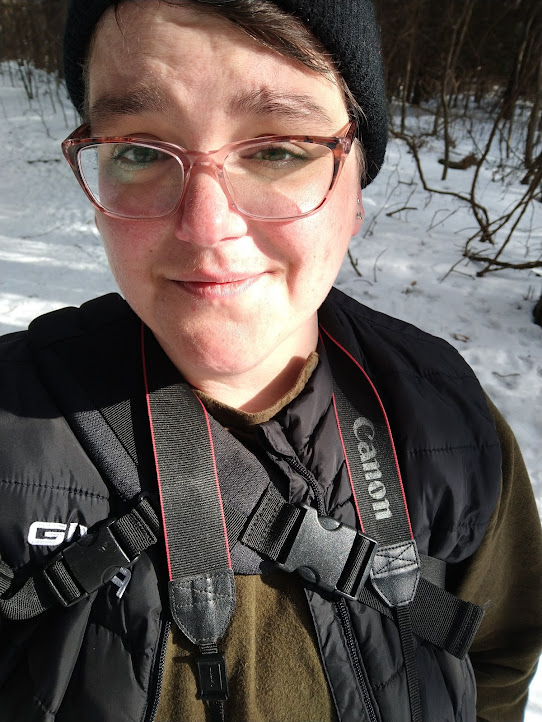 Lee regards the camera, squinting in bright light, Canon camera strap around their neck and snow visible in the background.